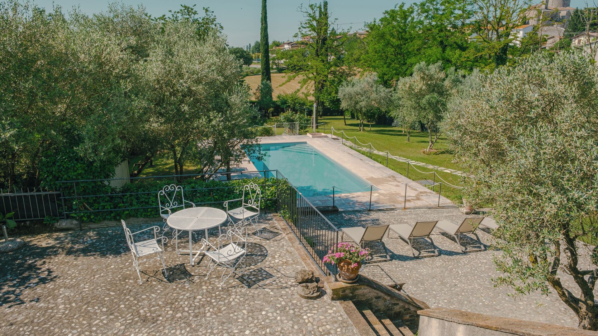 An outdoor swimming pool with lounge chairs and a table, surrounded by trees and a view of a village.