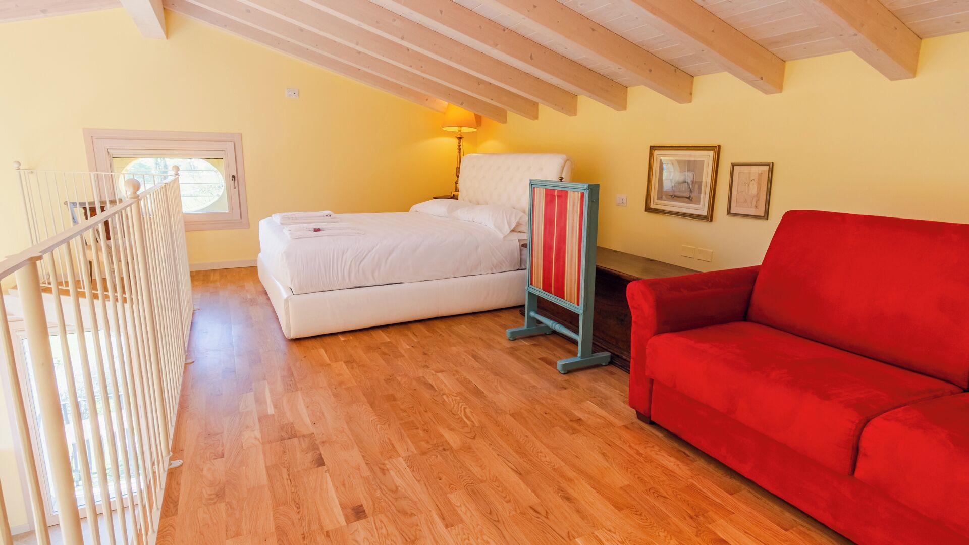 Une pièce avec un lit, un canapé rouge, un plancher en bois, une lampe et des tableaux encadrés accrochés au mur.