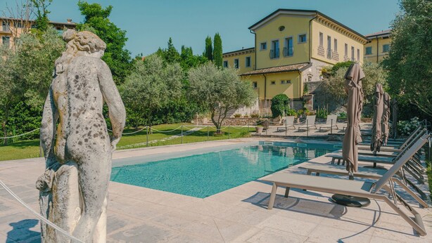 Zona de piscina con una estatua, sillones de descanso y un edificio al fondo.