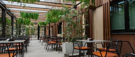 An indoor dining area with tables and chairs, surrounded by plants and wooden slats.