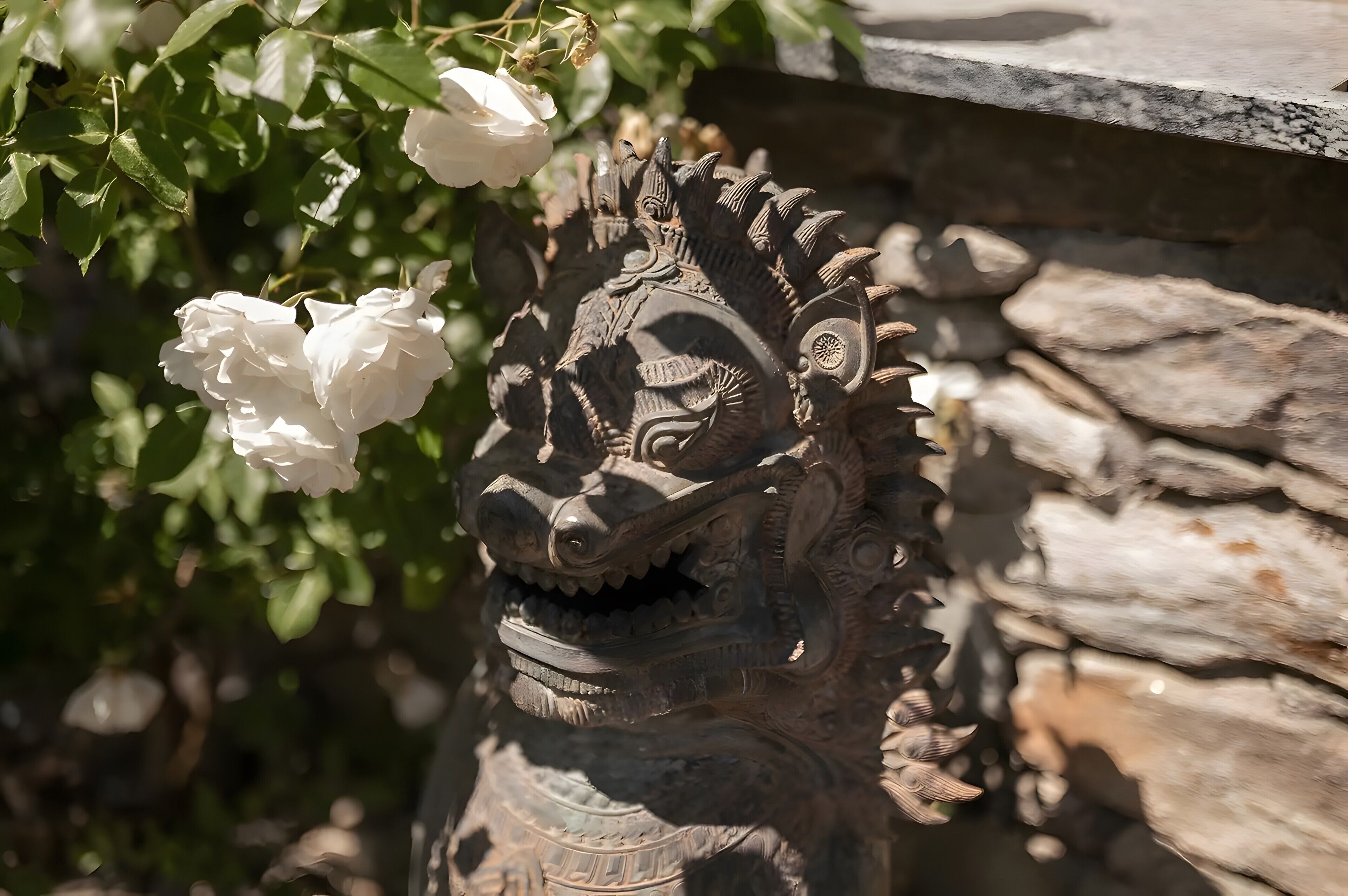 Une structure en pierre ornée d’une sculpture, entourée de verdure et de fleurs blanches.
