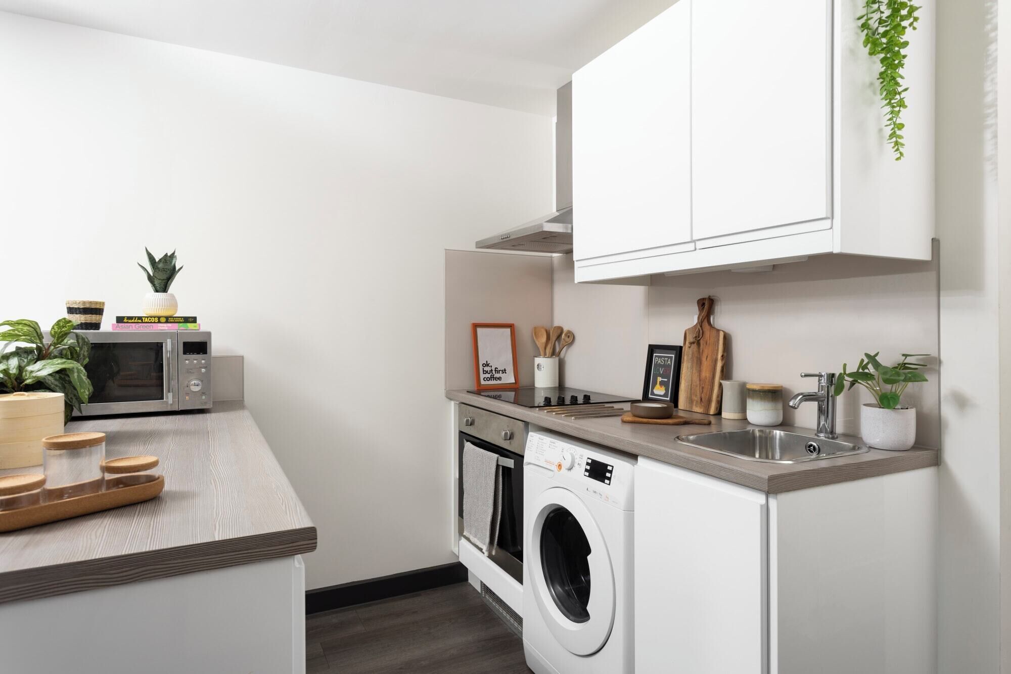 A compact kitchen with a microwave, oven, and washing machine, a sink, and a small countertop with a plant and a few items.