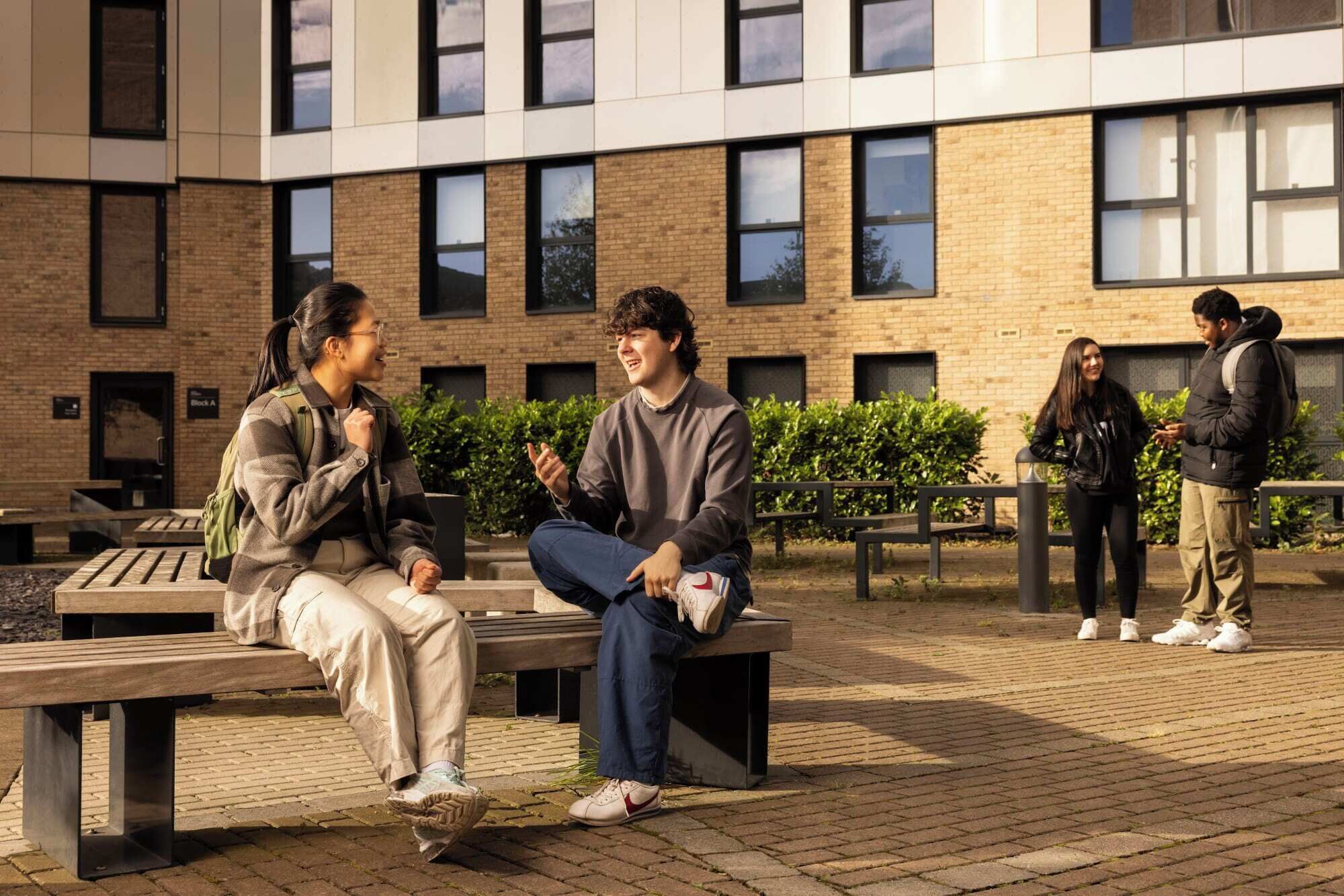A courtyard with a brick building, benches, and people sitting and standing.