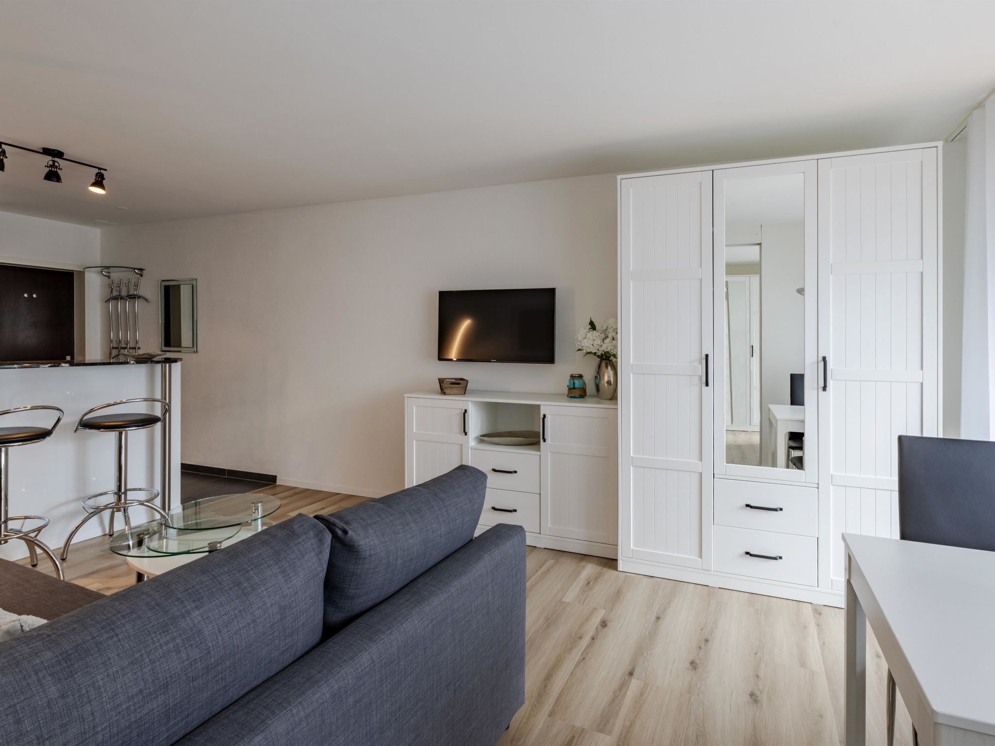 A modern living room with a grey sofa, a glass coffee table, and a TV mounted on the wall.