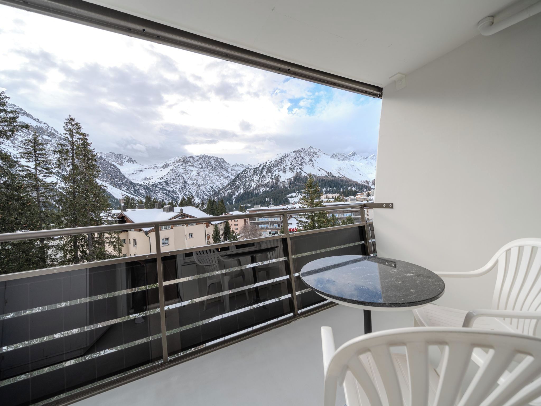 A balcony with a table and chairs, overlooking snow-covered mountains and buildings.