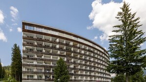 A multi-story hotel with a curved facade, balconies, and a parking area with cars. - Alexandra 513 Appartement/Fewo, Dusche, WC, 2-Bettwohnung by Interhome (Arosa)
