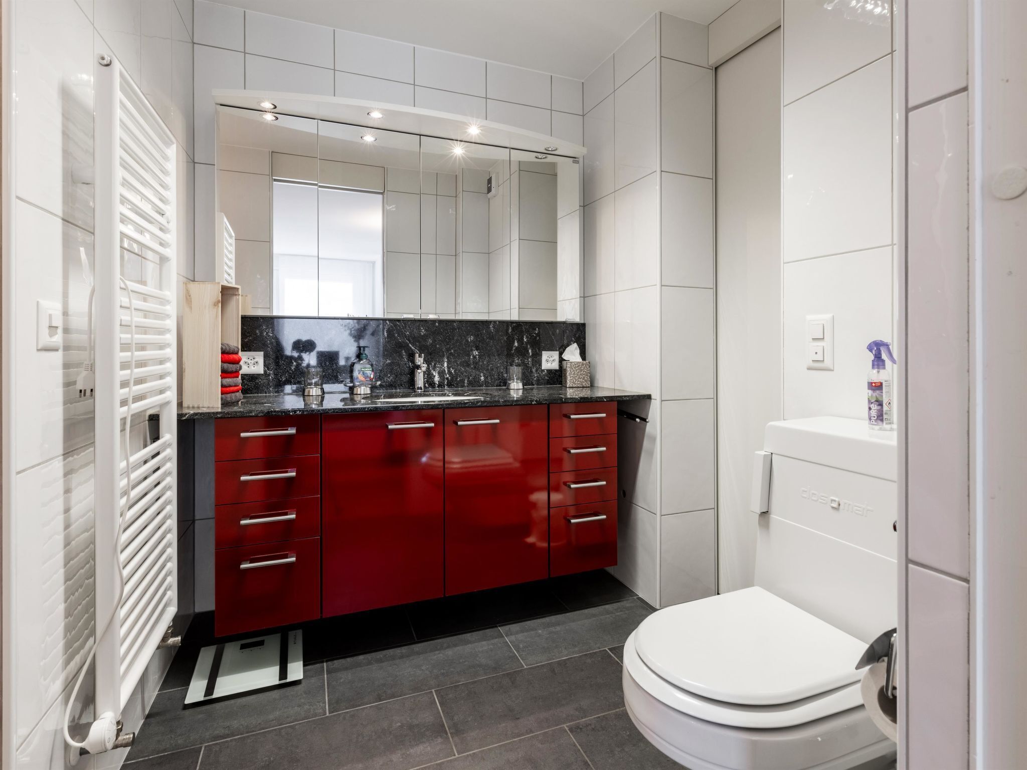 A modern bathroom with a white toilet, a red vanity, and a large mirror.