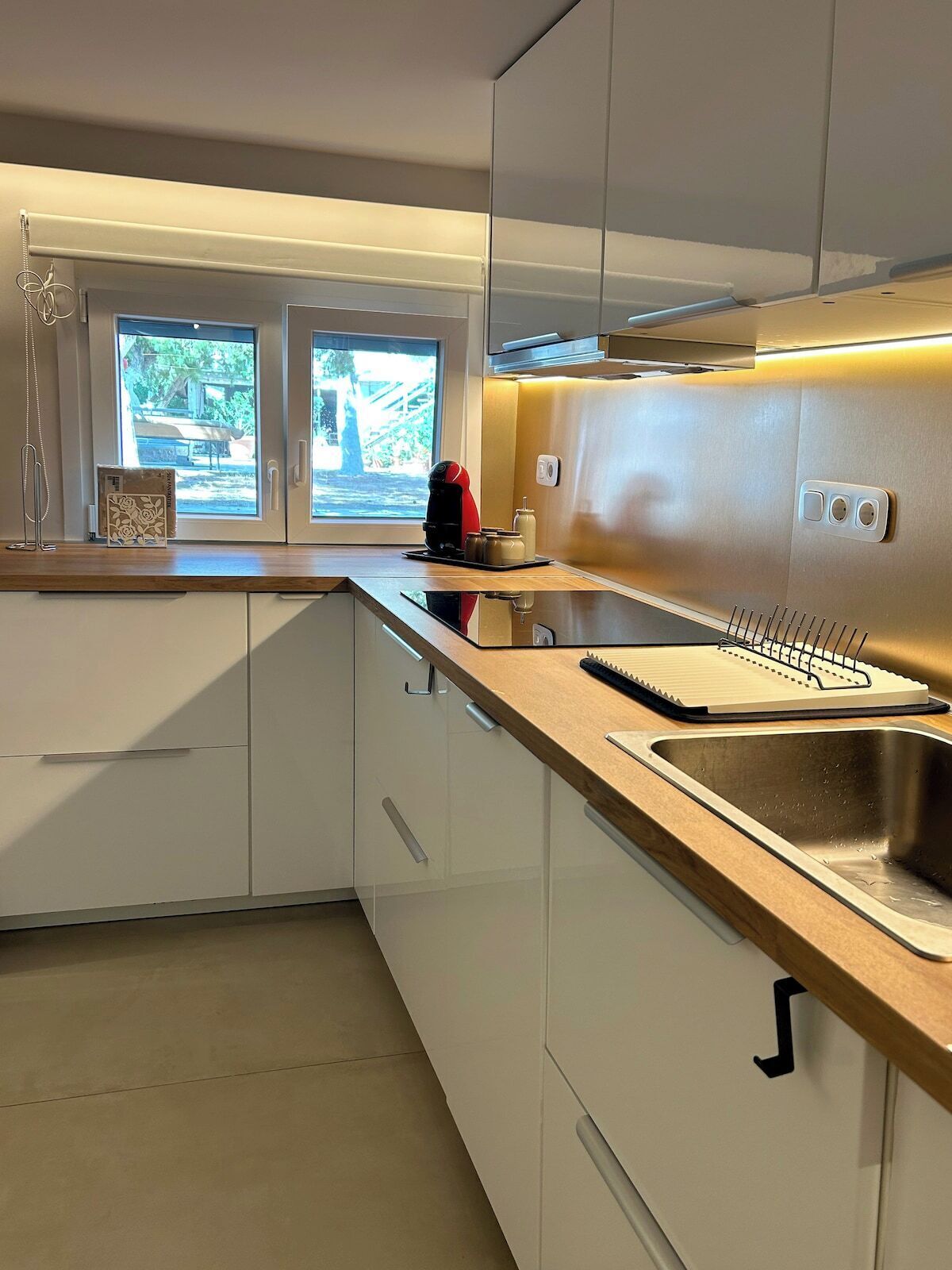 Une cuisine moderne avec des meubles blancs, un évier en acier inoxydable et une fenêtre donnant sur un paysage verdoyant.