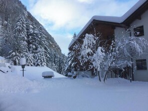 Ein schneebedecktes Gebäude mit geneigtem Dach, umgeben von schneebedeckten Bäumen und einer Bergkulisse.