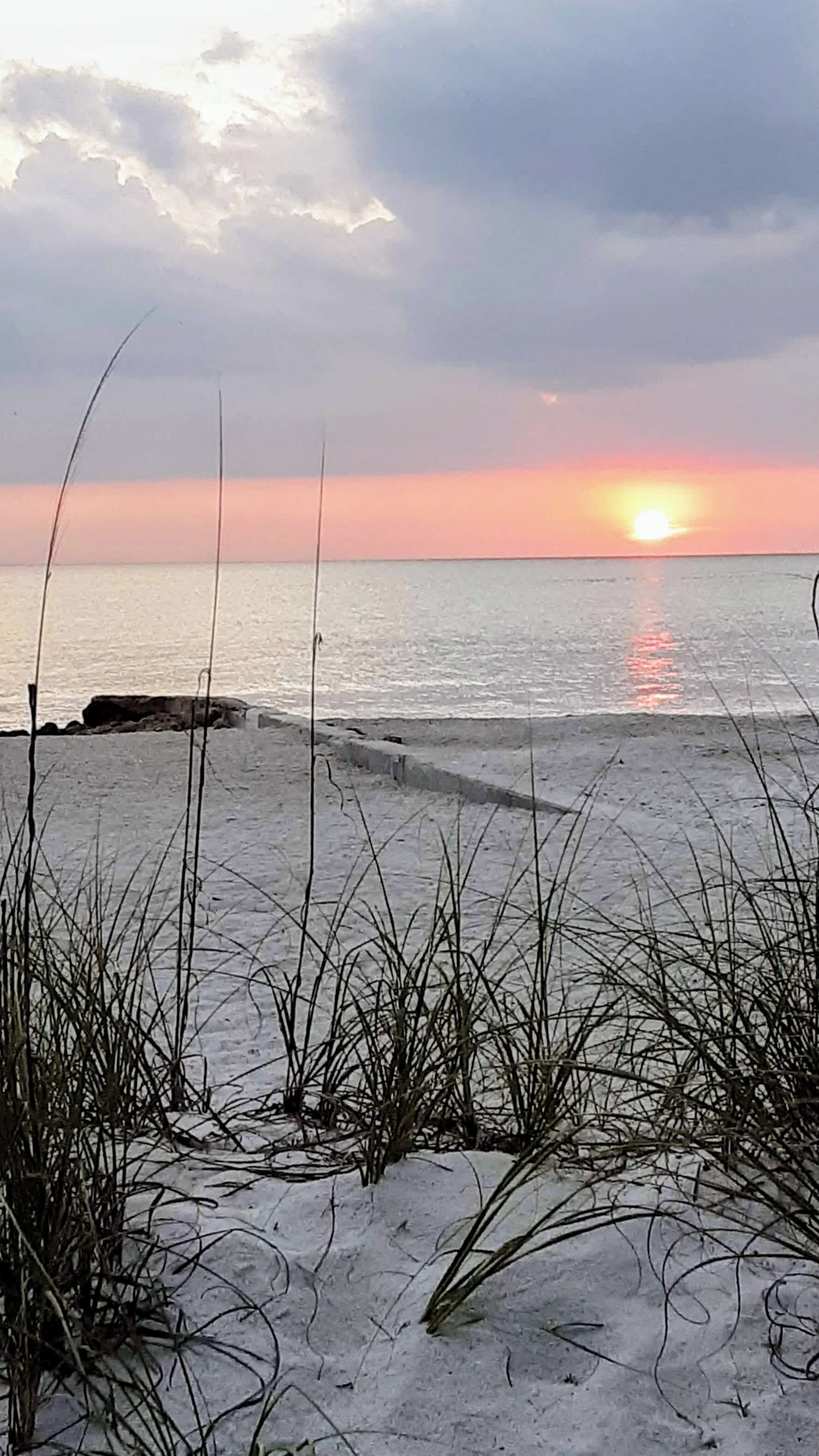 A beach with sand, grass, and a sunset over the water.