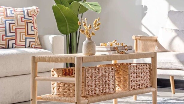 Ein Rattanzuhause mit gewebten Stauraumfächern, eine dekorative Vase mit getrocknetem Weizen und eine große grüne Pflanze im Hintergrund.