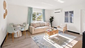 Ein Wohnzimmer mit Sofa, Esstisch und Stühlen, einem Couchtisch und einem großen Fenster mit Vorhängen.