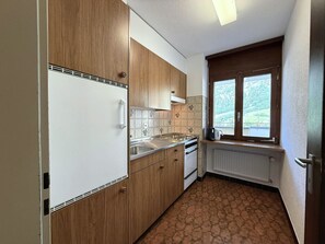 A compact kitchen with wooden cabinets, a white refrigerator, a built-in oven, and a window with a view of greenery. - Royal 51 # 4-bed apartment, Bad, WC 4-Bettwohnung, Bad, WC by Interhome (Leukerbad)