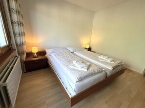 A double bed with white linens and towels, two bedside lamps, a wooden nightstand, a window with curtains, and a radiator. - Royal 51 # 4-bed apartment, Bad, WC 4-Bettwohnung, Bad, WC by Interhome (Leukerbad)