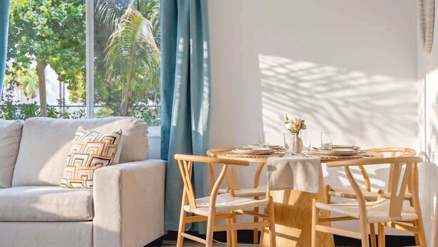 Ein Zimmer mit einer Couch, einem runden Holztisch mit Stühlen und einem Fenster mit Vorhängen.