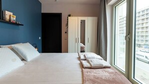 Une chambre d’hôtel avec un lit, une armoire, une fenêtre donnant sur des immeubles et une étagère remplie de livres.