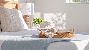 Een net opgemaakt bed met een witte deken, een bedlamp, een kleine potplant en een rieten dienblad met een theepot en kopjes op een witte tafel.