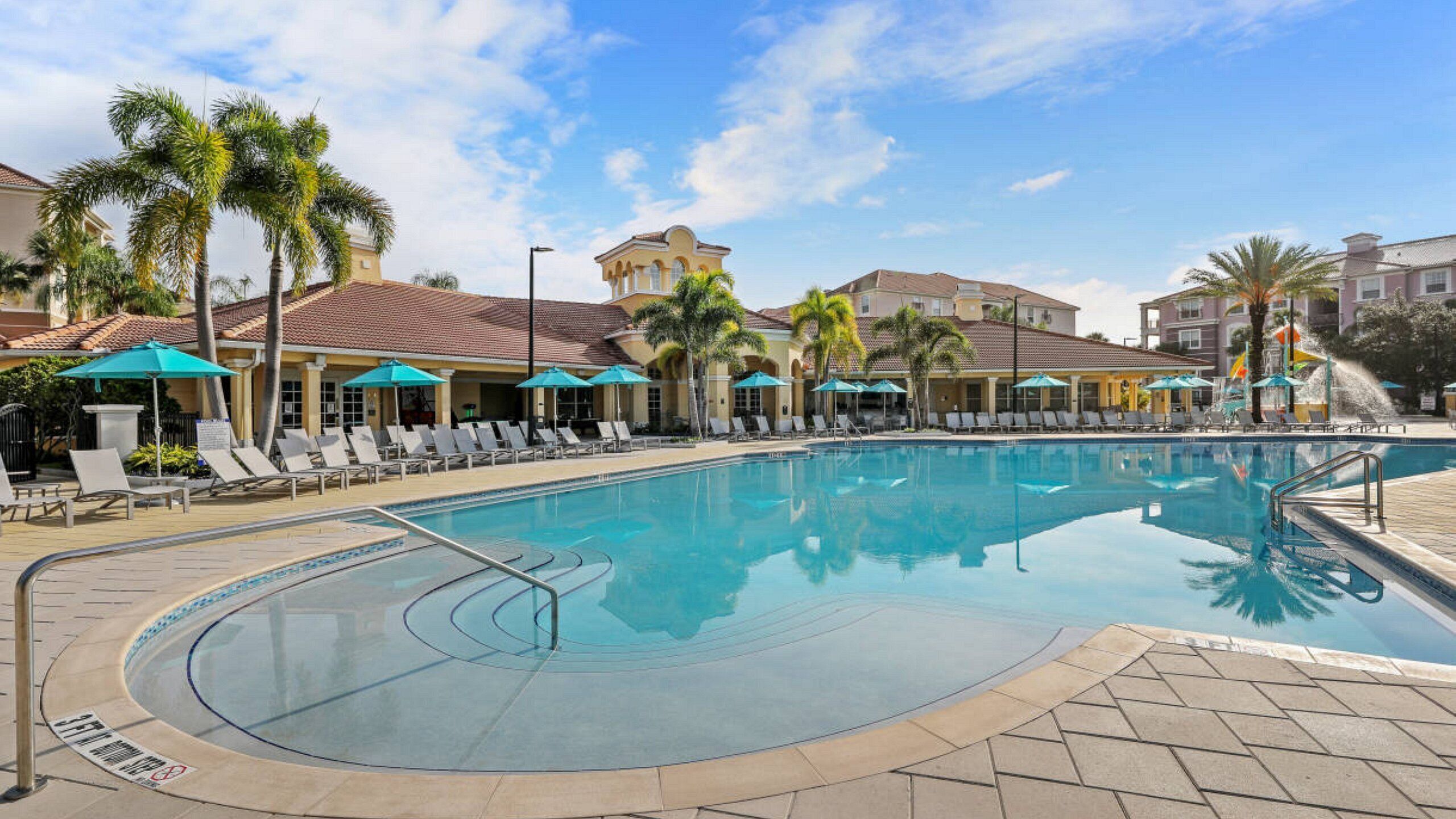A swimming pool with lounge chairs and umbrellas, surrounded by palm trees and buildings with a Mediterranean architectural style.