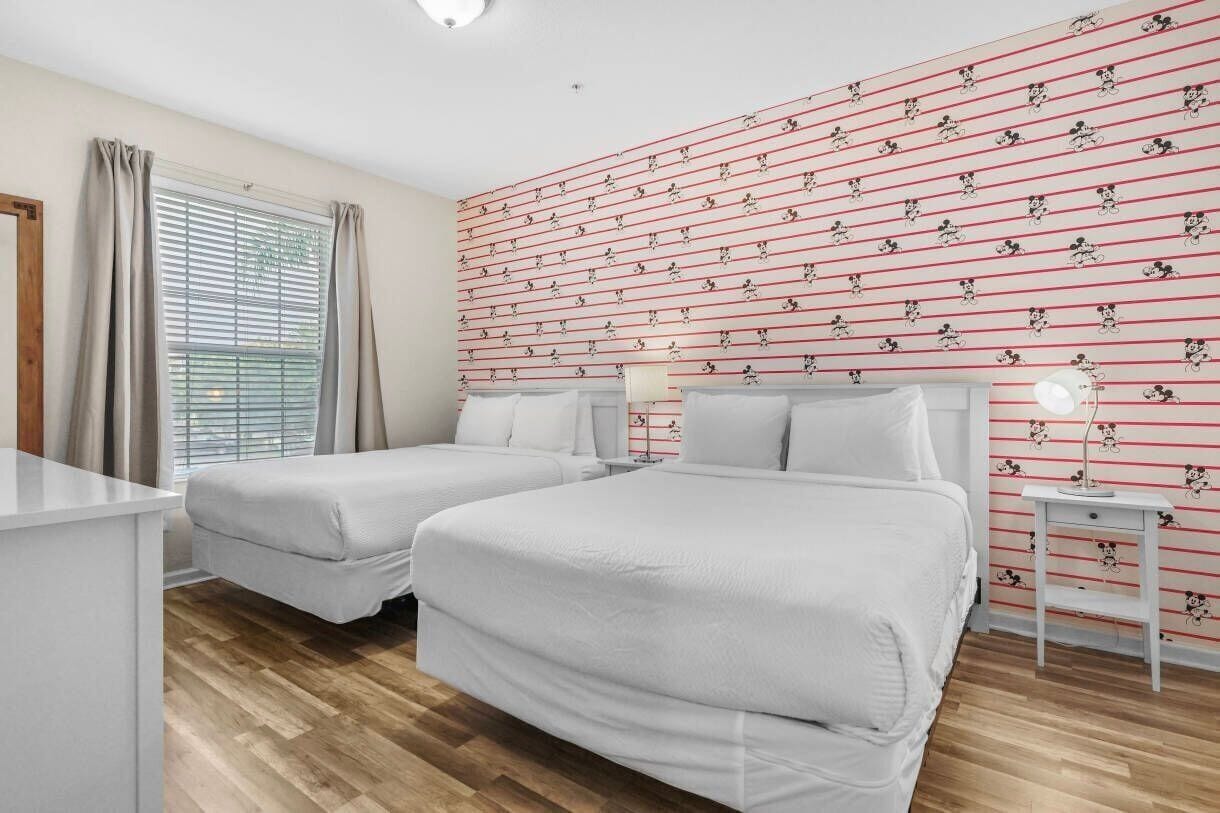 A room with two beds, a desk, a window with curtains, and a wall with a Mickey Mouse pattern.