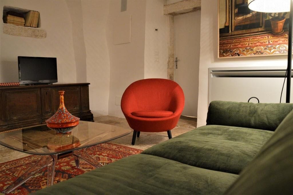 Ein Wohnzimmer mit einem grünen Sofa, einem roten Sessel, einem Glastisch mit einer Dekoration, einem Fernseher auf einer Holzkommode und einem gemusterten Teppich.
