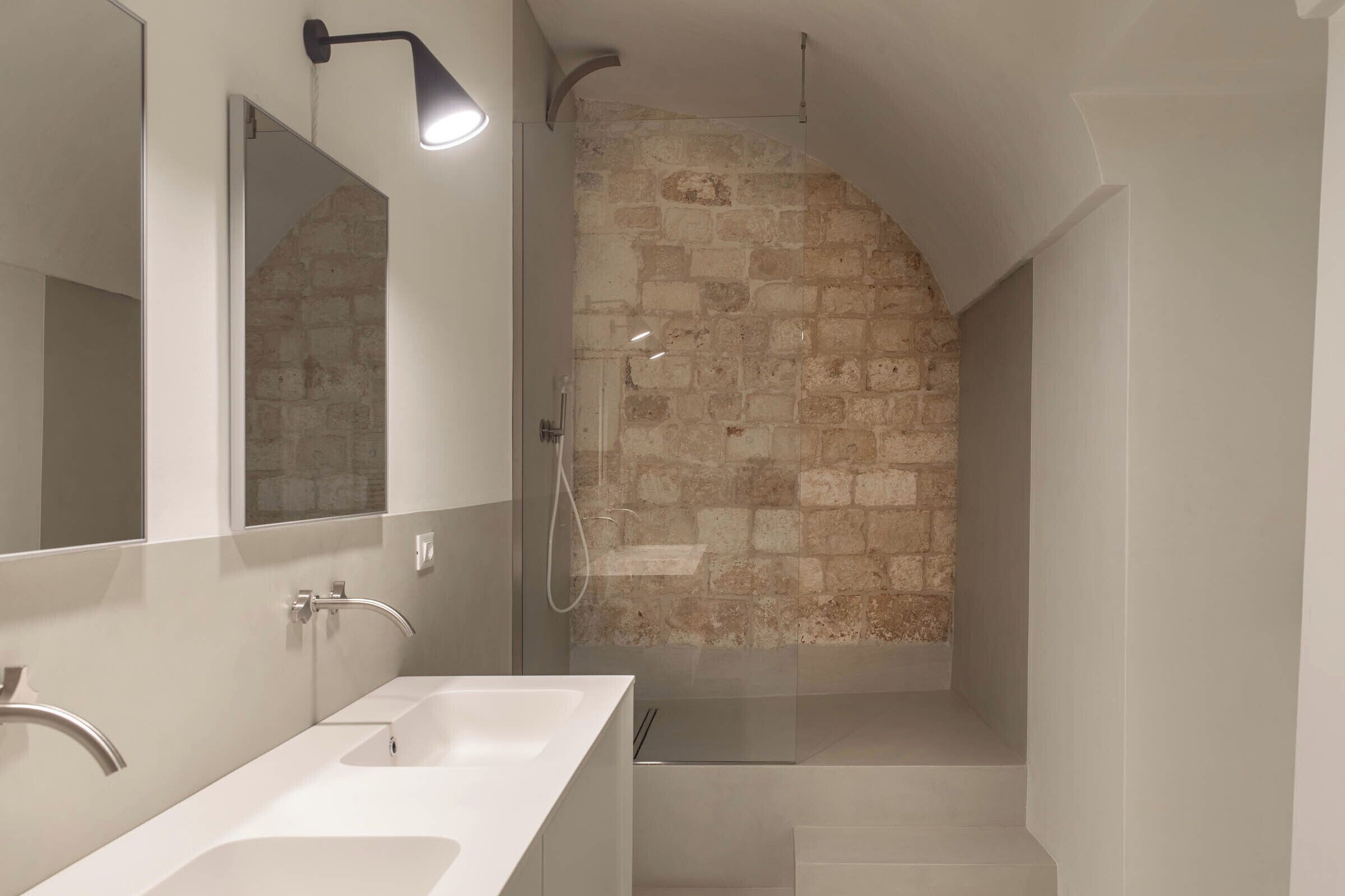 A modern bathroom with a large mirror, two sinks, a shower with a stone wall, and a light fixture.
