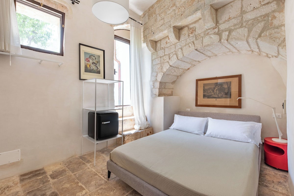 A bedroom with a stone wall, a bed with white bedding, a red side table, and a framed picture on the wall.