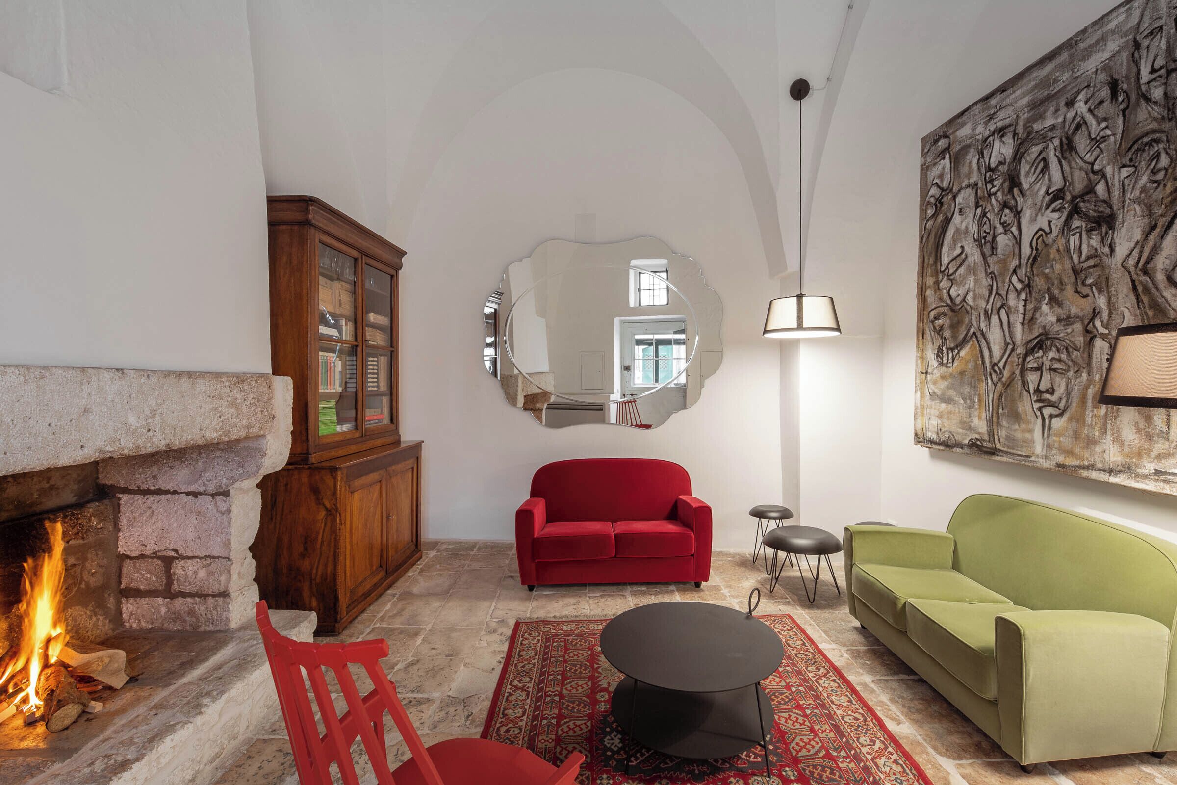 Un salon avec une cheminée, des canapés rouges et verts, une table ronde noire et un grand tableau abstrait accroché au mur.