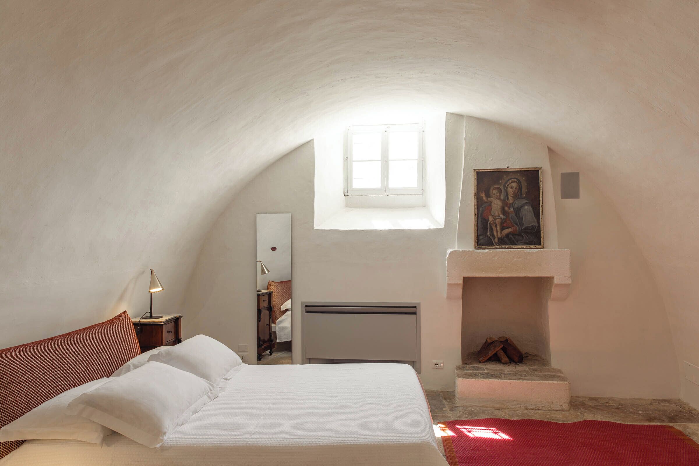 Een slaapkamer met een bed, nachtkastje, lamp, open haard en raam.