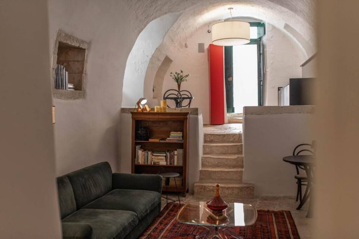 A cozy living room with a sofa, a bookshelf, a small table with a vase, and a staircase leading to an upper level.