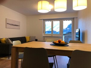 Een eetgedeelte met een houten tafel, stoelen, een fruitschaal, een bank met kussens en een raam met uitzicht op de bergen.