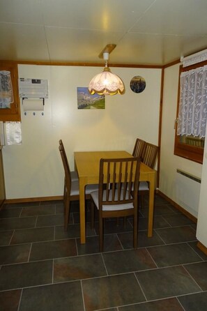 A dining area with a wooden table and chairs, a pendant light, a wall-mounted heater, and a window with a curtain. - Chalet Kondore 1 # 5-bed apartment, Dusche, WC 5-Bettwohnung, Dusche, WC by Interhome (Leukerbad)