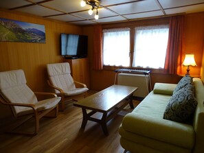 A room with a wooden floor, a sofa, two chairs, a table, a television, and a window with curtains. - Chalet Kondore 1 # 5-bed apartment, Dusche, WC 5-Bettwohnung, Dusche, WC by Interhome (Leukerbad)