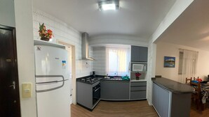 A compact kitchen with a refrigerator, stove, microwave, and a window with curtains. - Vacation rental apartment in the center of Canasvieiras. (Florianópolis)