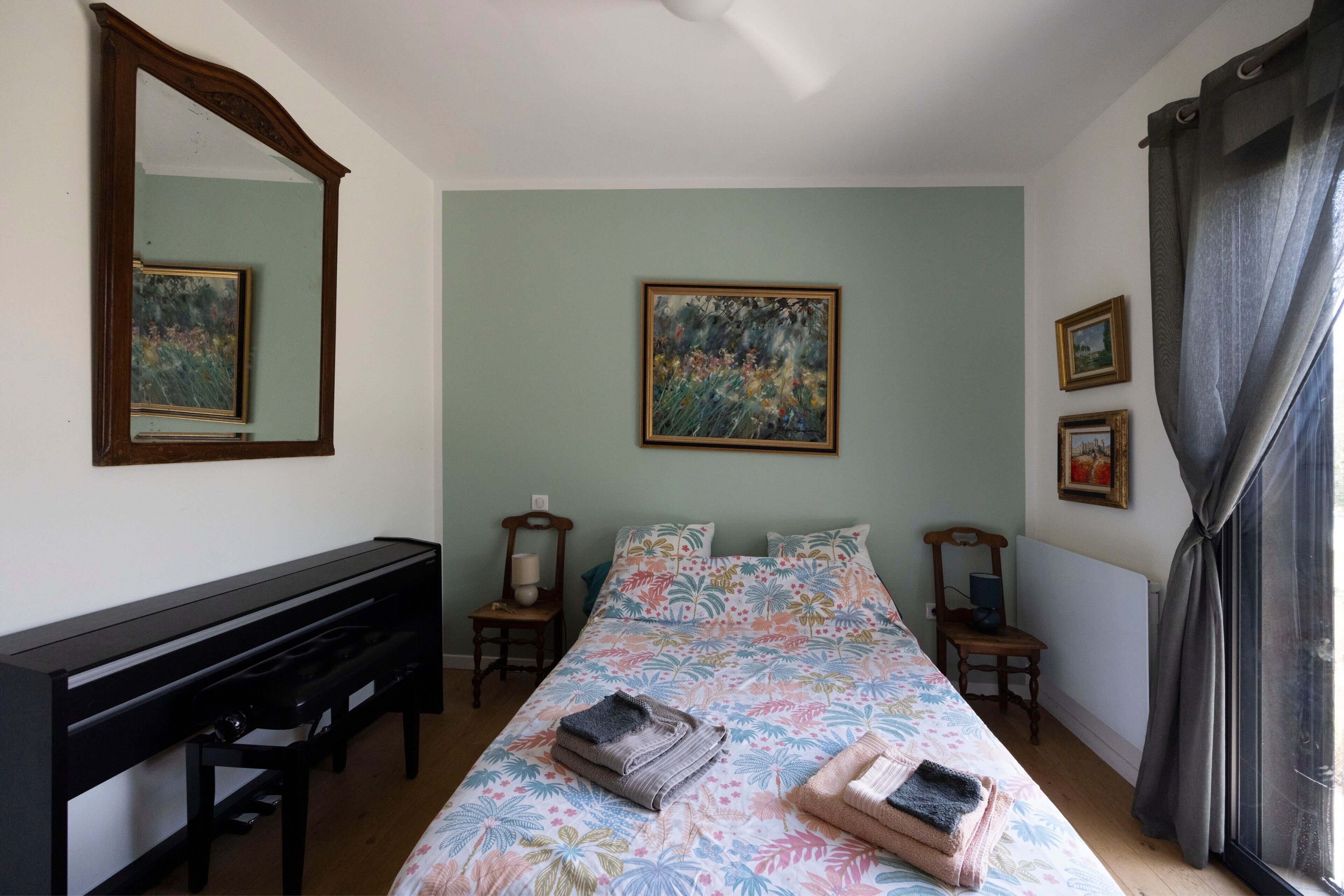 A bedroom with a bed, a desk, a chair, a mirror, and two paintings on the wall.