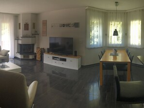 Ein modernes Wohnzimmer mit Flachbildfernseher, Kamin, Essbereich mit Tisch und Stühlen sowie großen Fenstern mit Vorhängen.