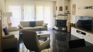 Ein modernes Wohnzimmer mit Sofa, Sesseln, einem Couchtisch, einem Flachbildfernseher, einem Kamin und einem großen Fenster mit transparenten Vorhängen.
