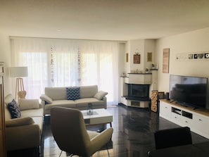 A modern living room with a sofa, armchairs, a coffee table, a flat-screen TV, a fireplace, and a large window with sheer curtains. - Edelweiss A 11 # 4-bed apartment, Dusche, WC 4-Bettwohnung, Dusche, WC by Interhome (Leukerbad)