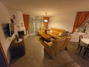 Ein Wohnzimmer mit Sofa, Sesseln, einem Couchtisch, einem Fernseher und einem Essbereich mit Tisch und Stühlen.