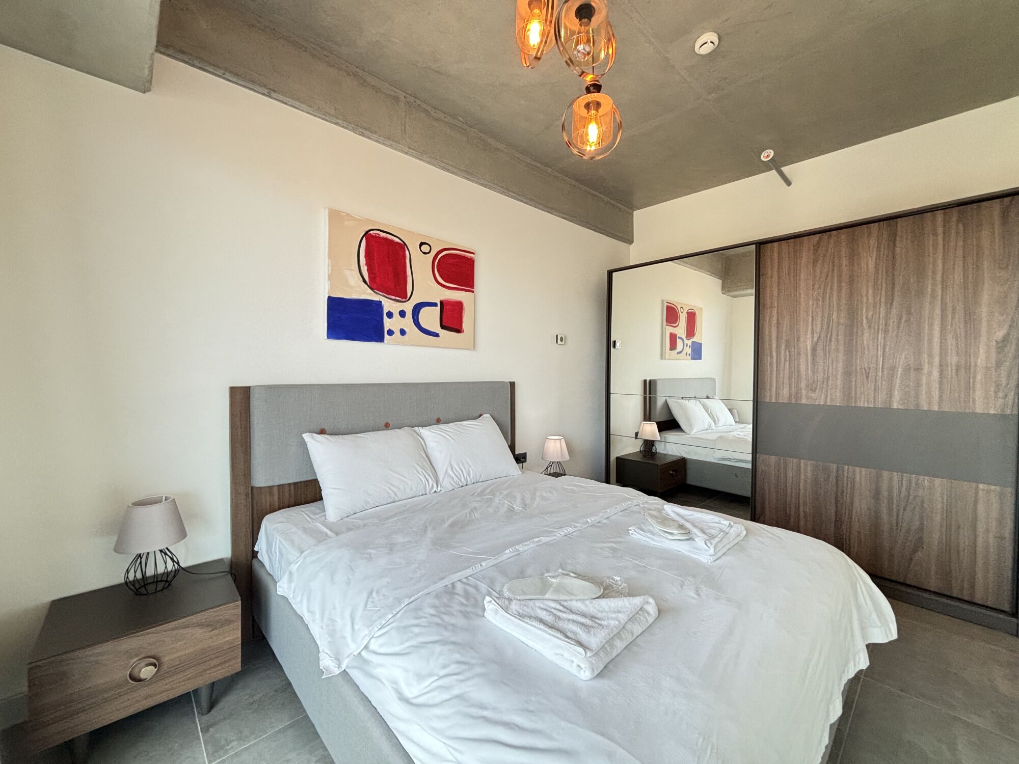Une chambre à coucher moderne avec un grand lit, des tables de chevet, une commode, un miroir et des œuvres d’art abstraites accrochées au mur.