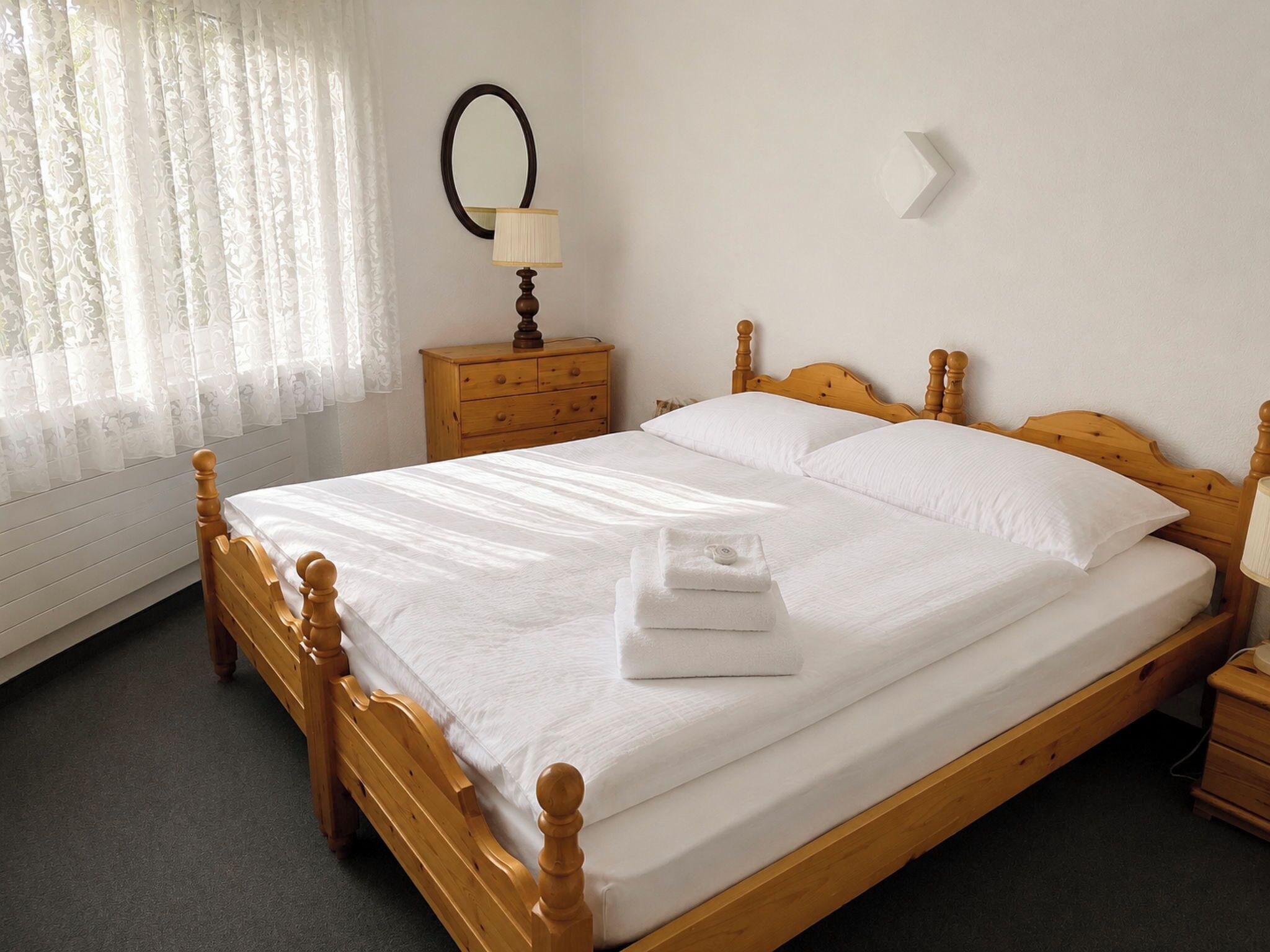 A wooden four-poster bed with white linens and a folded towel, a wooden nightstand with a lamp, and a window with sheer curtains.