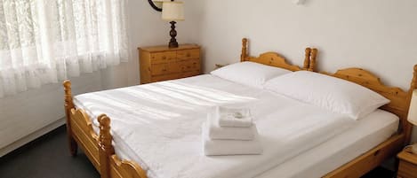 A wooden four-poster bed with white linens and a folded towel, a wooden nightstand with a lamp, and a window with sheer curtains.