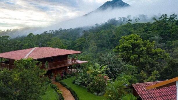 A wooden building with a red roof surrounded by lush greenery and a mountain in the background. - Costa Rican Paradise (La fortuna)