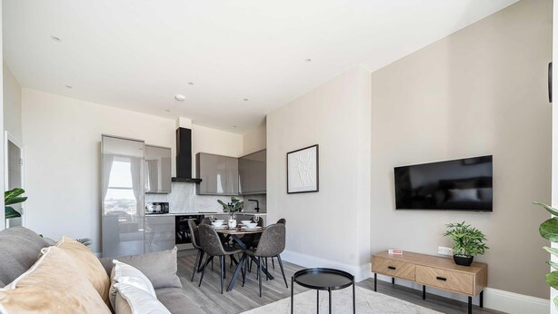 A modern living room with a sofa, a dining table with chairs, a kitchen area with cabinets, and a flat-screen TV mounted on the wall. - Shore Thing - Relaxing Sea View Getaway (Portsmouth)