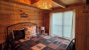 Ein Schlafzimmer einer Holzhütte mit einem Bett, einem 'Welcome'-Schild, einem Fenster mit Jalousien und einer Tür.
