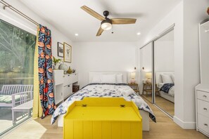 Ein Schlafzimmer mit einem Bett, einer gelben Bank, einem Deckenventilator, einem großen Spiegel und einem Fenster mit Vorhängen.
