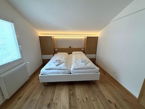 Un dormitorio con cama, cabecero de madera, paredes blancas y una ventana.