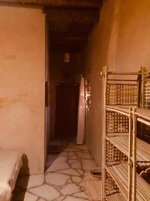 A narrow hallway with a stone floor, a bed on the left, and a metal shelf unit on the right. - tanirt ecolodge (Siwa)