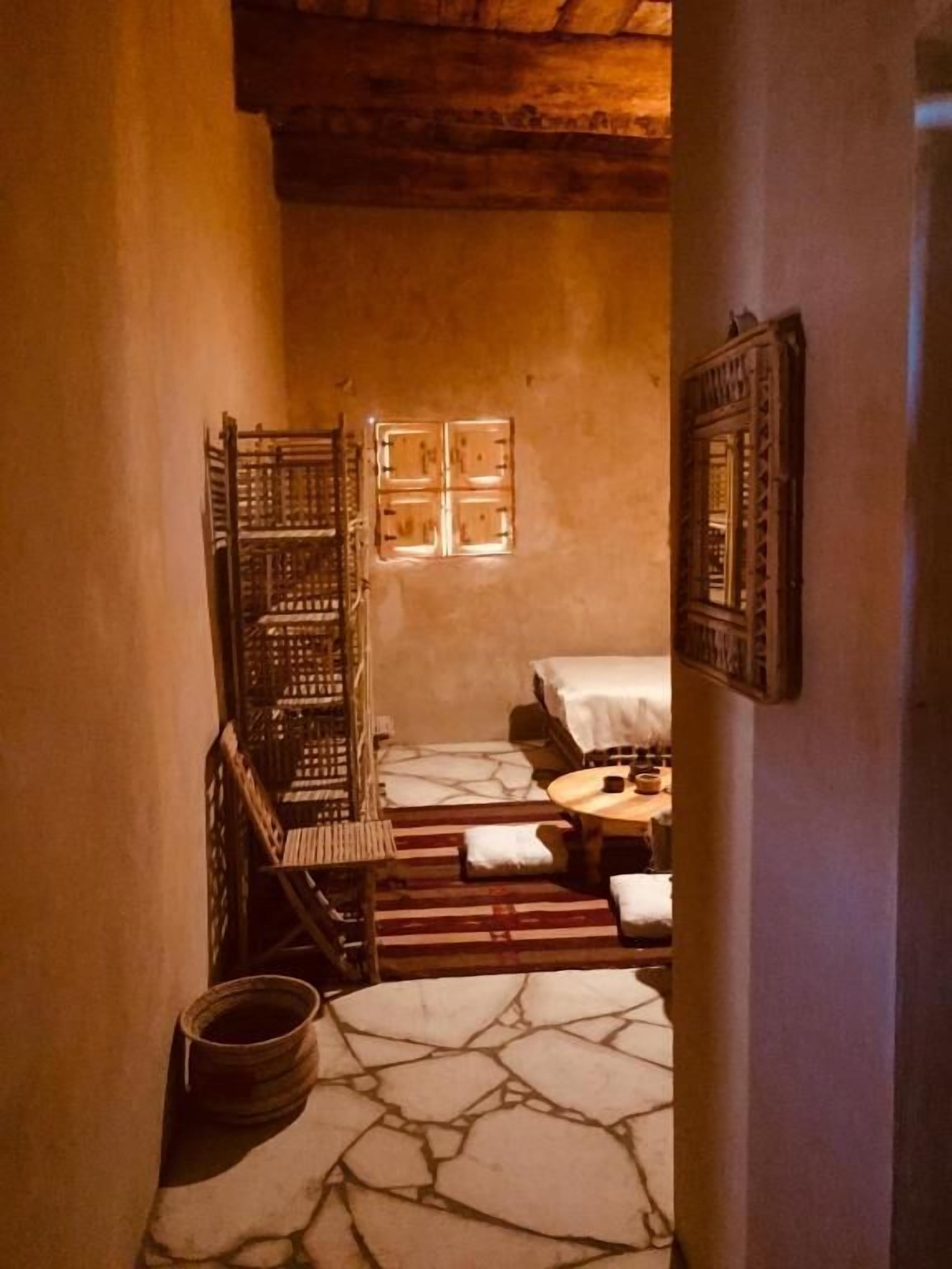 Un estrecho pasillo con suelo de piedra que conduce a una habitación con una cama, una silla de madera y una mesita.