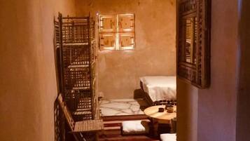 Un estrecho pasillo con suelo de piedra que conduce a una habitación con una cama, una silla de madera y una mesita.