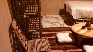 A stone-floored room with a woven chair, a small table with a candle, and a bed with a white cover. - tanirt ecolodge (Siwa)